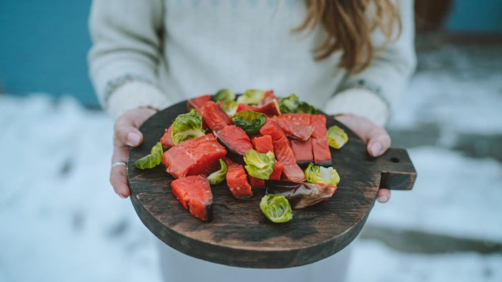Close-up of a person holding a wooden board with fresh salmon and Brussels sprouts on a snowy day.